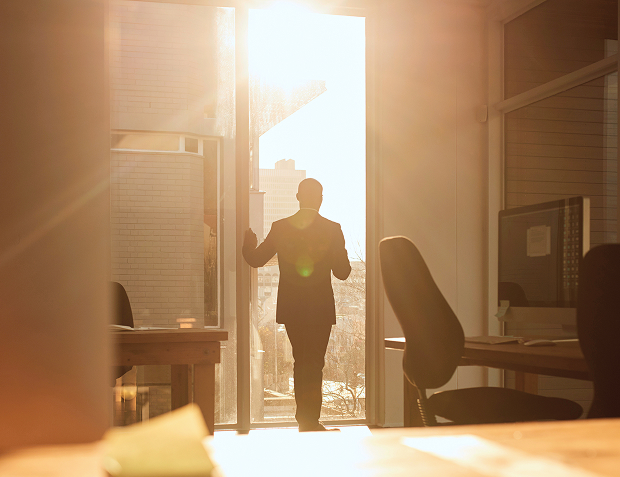 A man stands at a tall, narrow window, his office cast in the sun's glow.