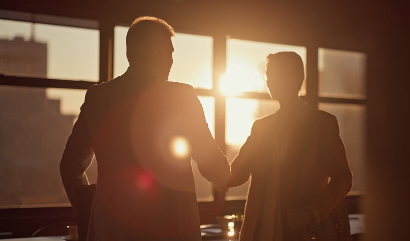 Two men in business attire, nearly in silhouette, shake hands inside an office with the sun setting through the window behind them.